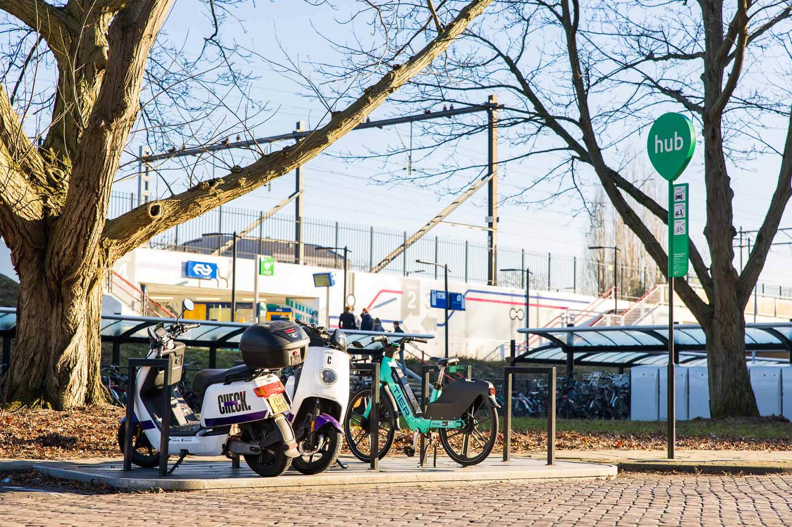 Foto van elektrische Check-scooters en een elektrische Tier-fiets aan een scooter- en motorrek bij een deelvervoerhub aan een treinstation.