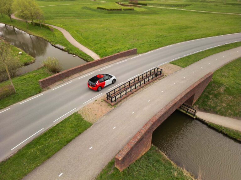 Een foto vanuit de lucht van een Greenwheels-auto die op een brug rijdt in een buitengebied met veel groen