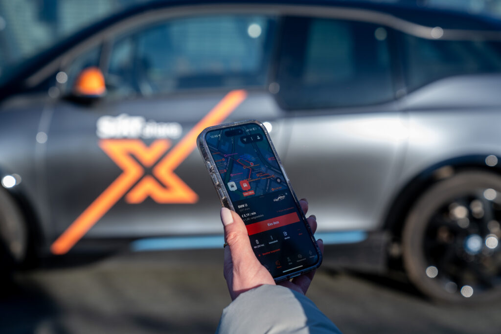 Een hand houdt een smartphone met een mobiliteitsapp omhoog, met op de achtergrond een geparkeerde deelauto met oranje merkaccenten.