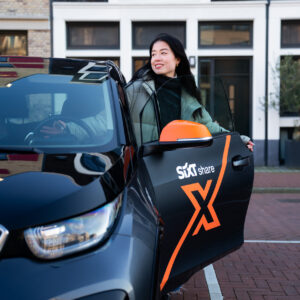 Een vrouw stapt glimlachend in een zwarte Sixt Share-deelauto met oranje accenten, terwijl de deur openstaat op een rustige parkeerplek in een stadse omgeving.