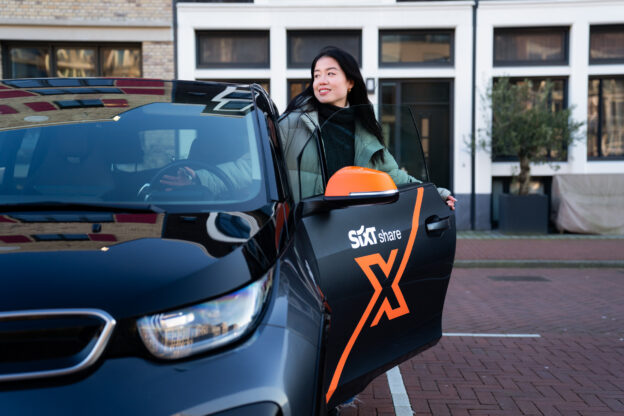 Een vrouw stapt glimlachend in een zwarte Sixt Share-deelauto met oranje accenten, terwijl de deur openstaat op een rustige parkeerplek in een stadse omgeving.