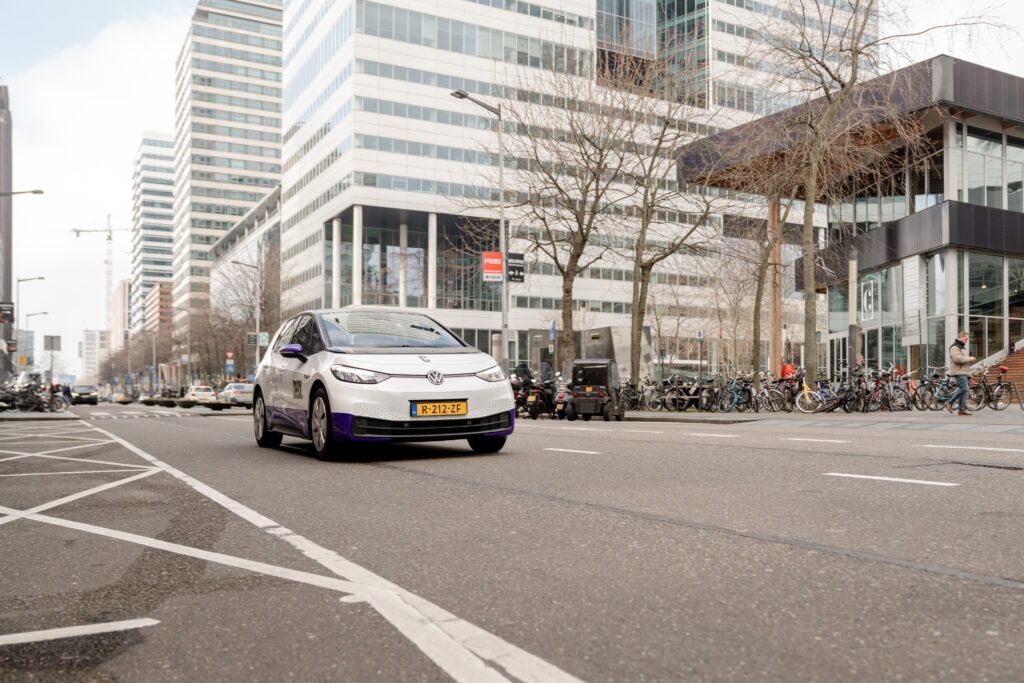 Elektrische deelauto rijdt door kantoorgebied in Nederlandse binnenstad; voorbeeld van deelmobiliteit in het dagelijks verkeer.