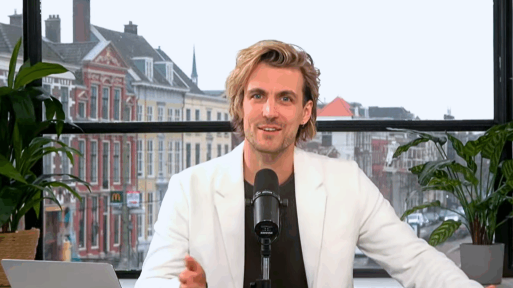 Still uit N!D Liveshow Modelbepalingen & Flexibele Parkeermachtiging: host in studio met microfoon; stadsgezicht op achtergrond