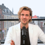 Still uit N!D Liveshow Modelbepalingen & Flexibele Parkeermachtiging: host in studio met microfoon; stadsgezicht op achtergrond