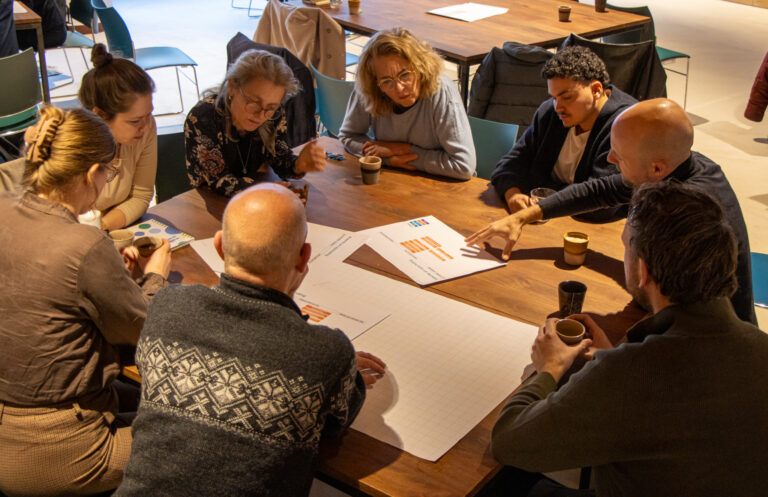 Een groep van acht mensen zit rond een tafel tijdens een werksessie, kijkend naar papieren met schema’s en samen pratend en wijzend terwijl ze overleggen.