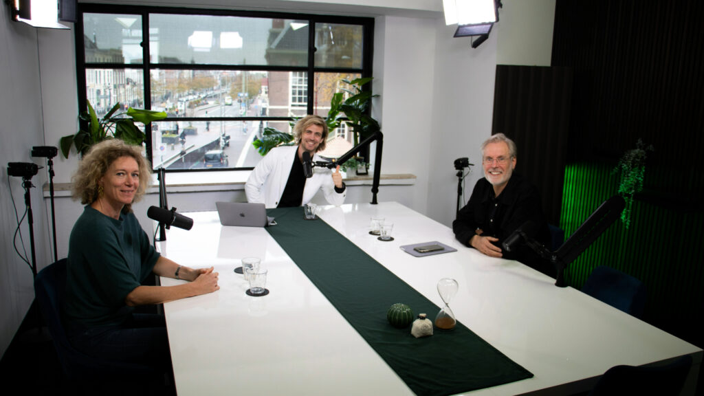 Drie mensen zitten aan een grote witte tafel in een studio-opstelling met microfoons en studiobelichting, klaar voor een podcast- of video-opname, met een raam op de achtergrond dat uitkijkt over een stadse straat.