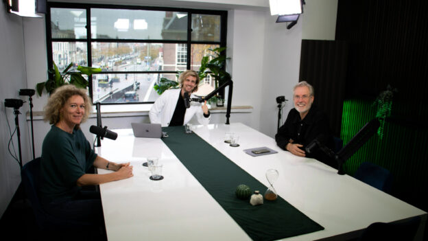 Drie mensen zitten aan een grote witte tafel in een studio-opstelling met microfoons en studiobelichting, klaar voor een podcast- of video-opname, met een raam op de achtergrond dat uitkijkt over een stadse straat.