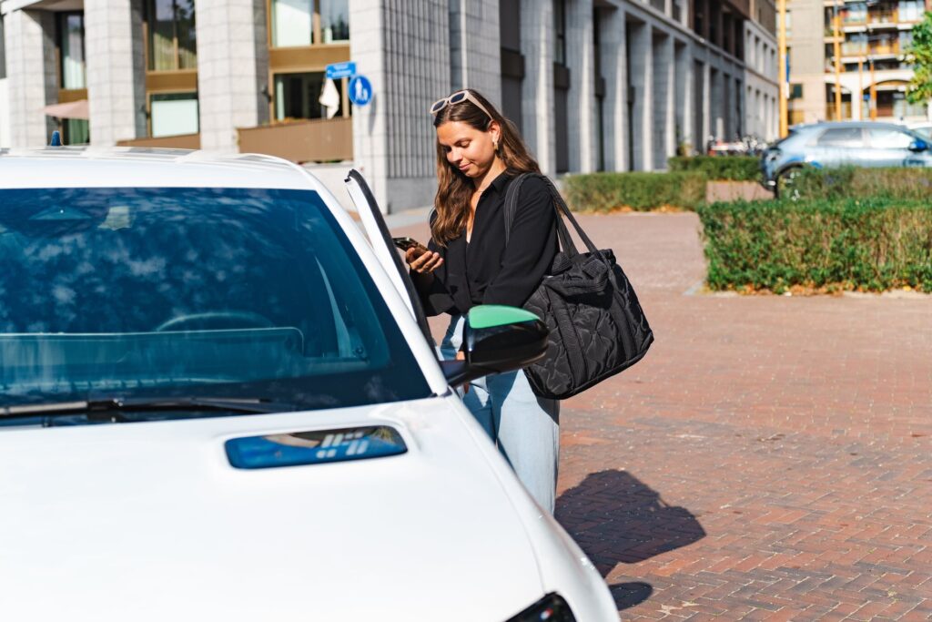 Een vrouw staat op straat naast een geparkeerde (deel)auto en kijkt op haar smartphone, terwijl ze de autodeur opent, met stedelijke bebouwing op de achtergrond.