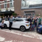 Groep bewoners poseert bij een witte deelauto in een straat, als start van een buurtinitiatief voor autodelen.