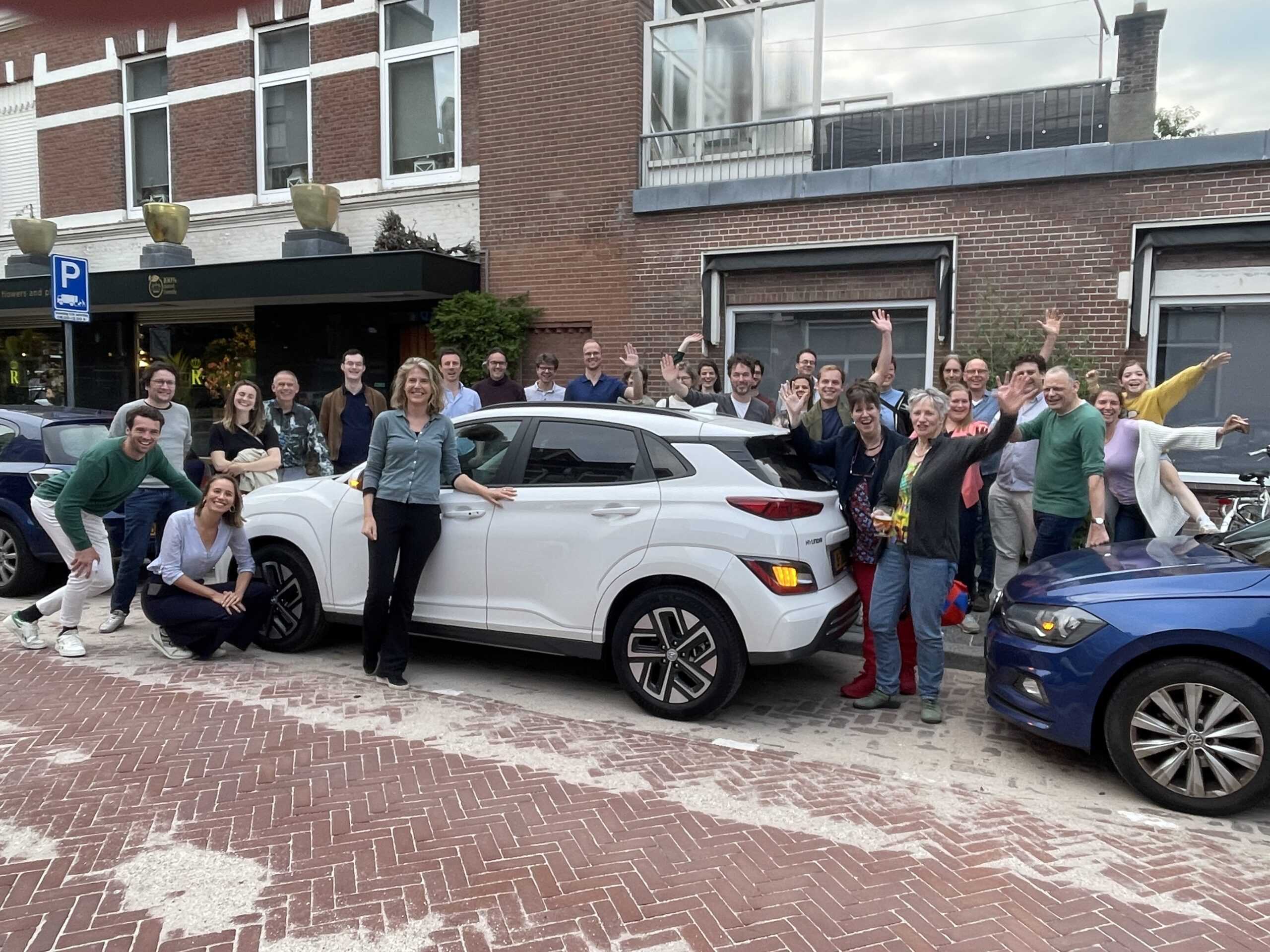Groep bewoners poseert bij een witte deelauto in een straat, als start van een buurtinitiatief voor autodelen.