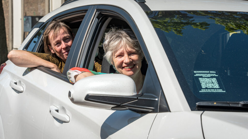 Twee vrouwen lachen vanuit een deelauto en kijken uit het open autoraam.