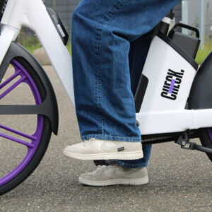 Close-up van iemands benen en voeten op de pedalen van een witte deelfiets met paarse wielen (Check), stilstaand op een verharde ondergrond.