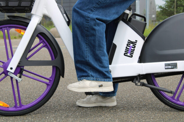 Close-up van iemands benen en voeten op de pedalen van een witte deelfiets met paarse wielen (Check), stilstaand op een verharde ondergrond.
