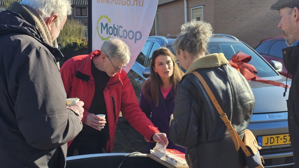 BuurtbewoGners in gesprek bij een informatiebijeenkomst over autodelen, bij een deelauto met een MobiGo-banner op de achtergrond.