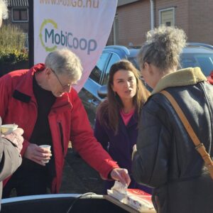 BuurtbewoGners in gesprek bij een informatiebijeenkomst over autodelen, bij een deelauto met een MobiGo-banner op de achtergrond.