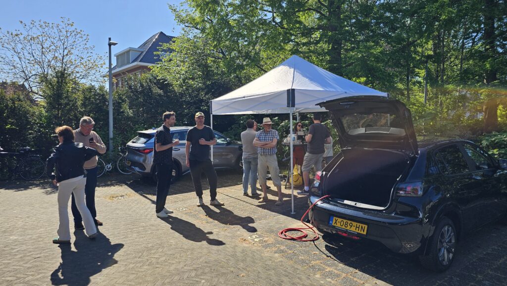 Buurtbijeenkomst rond deelmobiliteit op een zonnige dag, met bewoners bij een tent en een elektrische auto die wordt opgeladen op een parkeerplaats.